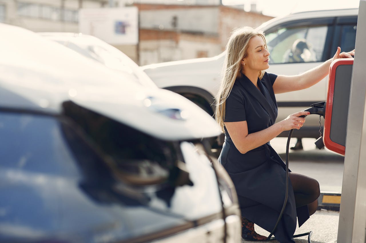 Side view of full length female driver in casual dress squatting near station while charging electric automobile for continuation of driving